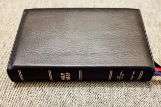 A black leather-bound book with the title 'Holy Bible' embossed in gold on the spine is lying flat on a textured surface. The book is thick, with several multi-colored ribbon markers peeking out from the bottom right corner.