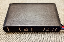 A black leather-bound book with the title 'Holy Bible' embossed in gold on the spine is lying flat on a textured surface. The book is thick, with several multi-colored ribbon markers peeking out from the bottom right corner.