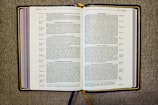 An open book displaying a passage from the Bible, specifically from the Gospel of Luke. The pages are filled with text in two columns, featuring headings such as 'A Warning Against Hypocrisy' and 'Do Not Worry.' The book rests on a textured fabric surface, and there are multiple ribbon bookmarks, including a prominent gold one.