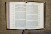 An open book displaying a passage from the Bible, specifically from the Gospel of Luke. The pages are filled with text in two columns, featuring headings such as 'A Warning Against Hypocrisy' and 'Do Not Worry.' The book rests on a textured fabric surface, and there are multiple ribbon bookmarks, including a prominent gold one.