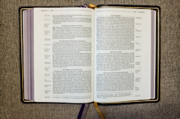 An open book displaying a passage from the Bible, specifically from the Gospel of Luke. The pages are filled with text in two columns, featuring headings such as 'A Warning Against Hypocrisy' and 'Do Not Worry.' The book rests on a textured fabric surface, and there are multiple ribbon bookmarks, including a prominent gold one.