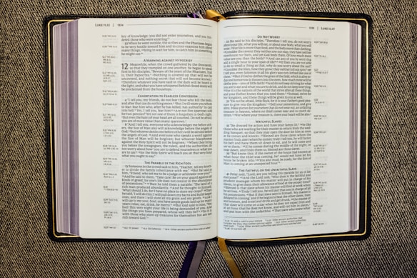 An open book displaying a passage from the Bible, specifically from the Gospel of Luke. The pages are filled with text in two columns, featuring headings such as 'A Warning Against Hypocrisy' and 'Do Not Worry.' The book rests on a textured fabric surface, and there are multiple ribbon bookmarks, including a prominent gold one.