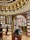 A luxurious library interior with an elegant design. The room features ornate wooden bookshelves filled with a variety of books, arranged in a circular pattern under a domed ceiling adorned with golden arches and intricate detailing. Warm lighting enhances the rich brown tones, and there are several round tables displaying books and electronic tablets in protective cases.