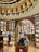 A luxurious library interior with an elegant design. The room features ornate wooden bookshelves filled with a variety of books, arranged in a circular pattern under a domed ceiling adorned with golden arches and intricate detailing. Warm lighting enhances the rich brown tones, and there are several round tables displaying books and electronic tablets in protective cases.
