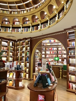 A luxurious library interior with an elegant design. The room features ornate wooden bookshelves filled with a variety of books, arranged in a circular pattern under a domed ceiling adorned with golden arches and intricate detailing. Warm lighting enhances the rich brown tones, and there are several round tables displaying books and electronic tablets in protective cases.