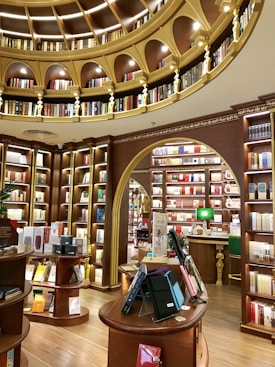 A luxurious library interior with an elegant design. The room features ornate wooden bookshelves filled with a variety of books, arranged in a circular pattern under a domed ceiling adorned with golden arches and intricate detailing. Warm lighting enhances the rich brown tones, and there are several round tables displaying books and electronic tablets in protective cases.