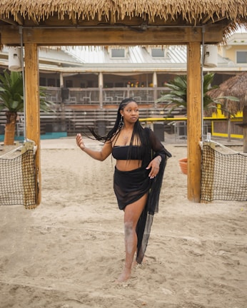 A woman in a black two-piece swimsuit and sheer cover-up stands on a sandy area. She is beneath a wooden structure with a thatched roof. In the background, there are tropical plants and a building with an open patio.
