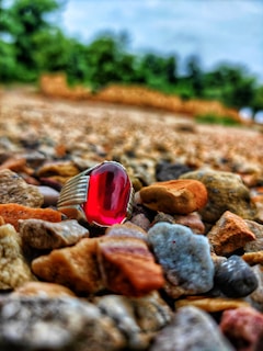 A silver ring with a vivid red gem is placed on a surface covered with small, multicolored pebbles. The backdrop consists of an out-of-focus natural setting with greenery, which gives a sense of depth to the image.