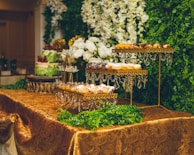 An elegant dessert table display features tiered trays adorned with a variety of pastries and sweets, all set against a backdrop of lush greenery and white floral arrangements. The table is covered with an ornate gold tablecloth, and crystal decorations hang from the trays.