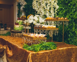 An elegant dessert table display features tiered trays adorned with a variety of pastries and sweets, all set against a backdrop of lush greenery and white floral arrangements. The table is covered with an ornate gold tablecloth, and crystal decorations hang from the trays.