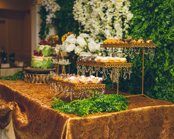 An elegant dessert table display features tiered trays adorned with a variety of pastries and sweets, all set against a backdrop of lush greenery and white floral arrangements. The table is covered with an ornate gold tablecloth, and crystal decorations hang from the trays.