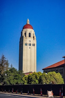 A tall, beige tower with a red dome stands prominently against a clear blue sky. Surrounding the base of the tower are green trees and red-roofed buildings. A black fence lines the foreground, with several orange traffic cones and a sign indicating 'Uneven Pavement Surface'.