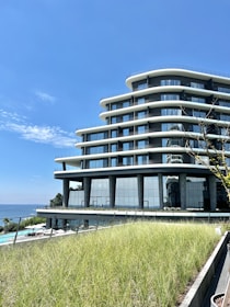 A modern, multi-story building with curvilinear architecture set against a bright blue sky. The facade features extensive use of glass, reflecting the sky and surroundings. In the foreground, a lush, green rooftop garden adds a natural element. The ocean can be seen in the background, enhancing the scenic view.