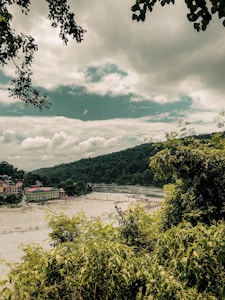 A lush green landscape with dense foliage in the foreground and a river cutting across the scene. People gather along the riverbank on a sandy shore near colorful buildings. A backdrop of forested hills rises beneath a cloudy sky.