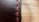 A close-up view of a black leather surface with prominent red stitching running vertically. The texture of the leather is visible, with fine lines and slight imperfections giving it a rugged look. The background is softly blurred, creating a warm contrast with the leather's dark hue.