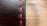A close-up view of a black leather surface with prominent red stitching running vertically. The texture of the leather is visible, with fine lines and slight imperfections giving it a rugged look. The background is softly blurred, creating a warm contrast with the leather's dark hue.
