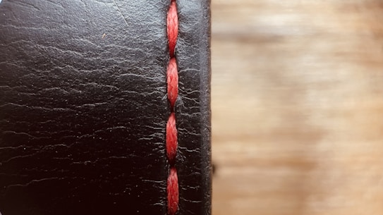 A close-up view of a black leather surface with prominent red stitching running vertically. The texture of the leather is visible, with fine lines and slight imperfections giving it a rugged look. The background is softly blurred, creating a warm contrast with the leather's dark hue.