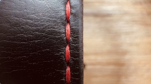 A close-up view of a black leather surface with prominent red stitching running vertically. The texture of the leather is visible, with fine lines and slight imperfections giving it a rugged look. The background is softly blurred, creating a warm contrast with the leather's dark hue.