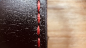 A close-up view of a black leather surface with prominent red stitching running vertically. The texture of the leather is visible, with fine lines and slight imperfections giving it a rugged look. The background is softly blurred, creating a warm contrast with the leather's dark hue.
