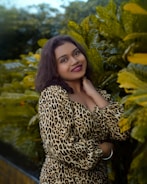 A woman with dark hair smiles while standing in front of lush green foliage. She wears a leopard print outfit and has her hand resting near her neck.