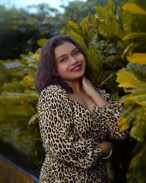 A woman with dark hair smiles while standing in front of lush green foliage. She wears a leopard print outfit and has her hand resting near her neck.