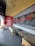 A modern fast-food restaurant interior features wooden ceiling slats, red and black decor, and seating arrangements with beige chairs and black tables. Red neon signage and a decorative wall display add to the ambiance.