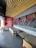 A modern fast-food restaurant interior features wooden ceiling slats, red and black decor, and seating arrangements with beige chairs and black tables. Red neon signage and a decorative wall display add to the ambiance.