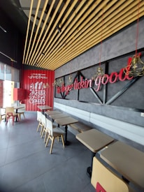 A modern fast-food restaurant interior features wooden ceiling slats, red and black decor, and seating arrangements with beige chairs and black tables. Red neon signage and a decorative wall display add to the ambiance.