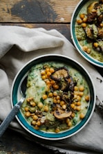 A close-up view of a bowl filled with creamy soup garnished with chickpeas, sautéed mushrooms, and green herbs. The soup has a rich and thick consistency with visible spinach leaves and pesto. The bowl is placed on a rustic wooden surface next to a light-colored cloth napkin.