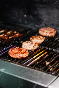 Four hamburger patties are being grilled on a black barbecue grill with visible grill marks. The patties appear juicy and are positioned in a row. The heat from the grill is causing some smoke and slight charring.
