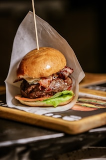 A well-cooked burger is presented on a wooden tray. The burger features a glossy bun, a juicy meat patty, crispy bacon strips, fresh lettuce, and a slice of tomato. The burger is held together with a wooden skewer and partially wrapped in white parchment paper.