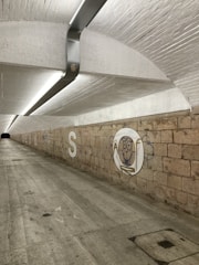 A long, underground tunnel featuring a curved ceiling lined with a fluorescent light. The walls are made of exposed brick, with graffiti art including a cartoonish face inside a circle and the letter 'S' prominently displayed. The floor is made of smooth concrete, with some scattered paint marks.
