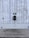 A modern electric vehicle charging station is mounted on a vertical concrete wall. The station is sleek and black, with a connected charging cable hanging down. The concrete wall has vertical grooves and is slightly weathered. The ground is paved with cobblestones, and there are small circular vents on the wall.