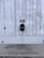 A modern electric vehicle charging station is mounted on a vertical concrete wall. The station is sleek and black, with a connected charging cable hanging down. The concrete wall has vertical grooves and is slightly weathered. The ground is paved with cobblestones, and there are small circular vents on the wall.