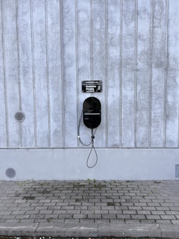 A modern electric vehicle charging station is mounted on a vertical concrete wall. The station is sleek and black, with a connected charging cable hanging down. The concrete wall has vertical grooves and is slightly weathered. The ground is paved with cobblestones, and there are small circular vents on the wall.