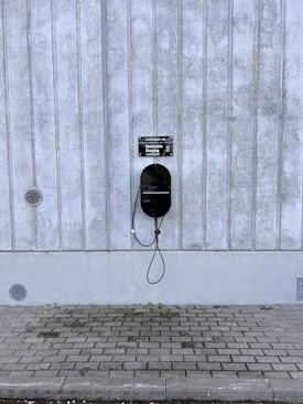 A modern electric vehicle charging station is mounted on a vertical concrete wall. The station is sleek and black, with a connected charging cable hanging down. The concrete wall has vertical grooves and is slightly weathered. The ground is paved with cobblestones, and there are small circular vents on the wall.