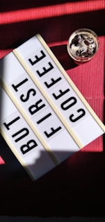 A lightbox with changeable letters spells out 'But First Coffee,' placed on a red textured surface. Beside it, a glass filled with coffee and cream, topped with dark chocolate shavings or similar garnishes, is seen. The setup is partially illuminated by natural light, creating a cozy atmosphere.