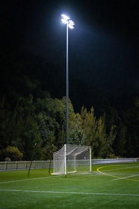A soccer goal post is illuminated by bright stadium lights on a field at night. The surroundings include a fence and dense trees in the background, creating a serene and quiet atmosphere.