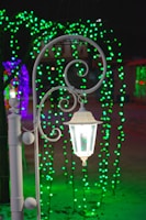 A white decorative street lamp is the focal point, emitting a soft glow against a background of vibrant green and purple string lights. These lights create an enchanting atmosphere, casting colorful reflections on the snowy ground.