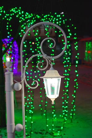 A white decorative street lamp is the focal point, emitting a soft glow against a background of vibrant green and purple string lights. These lights create an enchanting atmosphere, casting colorful reflections on the snowy ground.