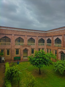 A courtyard with a well-maintained garden surrounded by a brick building featuring arched windows. The sky is overcast, suggesting an impending rain. The grassy area is lush, with trimmed hedges and a central tree providing a fresh, inviting look.
