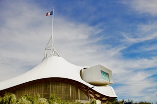 A modern architectural structure with a white, tent-like roof and a French flag on top. The sky is bright blue with scattered clouds. The building has a geometric extension with large windows and is surrounded by greenery.