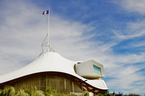 A modern architectural structure with a white, tent-like roof and a French flag on top. The sky is bright blue with scattered clouds. The building has a geometric extension with large windows and is surrounded by greenery.