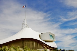 A modern architectural structure with a white, tent-like roof and a French flag on top. The sky is bright blue with scattered clouds. The building has a geometric extension with large windows and is surrounded by greenery.