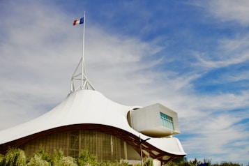 A modern architectural structure with a white, tent-like roof and a French flag on top. The sky is bright blue with scattered clouds. The building has a geometric extension with large windows and is surrounded by greenery.