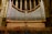 A large pipe organ is situated inside a cathedral, featuring numerous silver pipes arranged symmetrically against a backdrop of stained glass windows and ornate stone columns. The seating area in front of the organ is made up of wooden chairs.