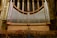 A large pipe organ is situated inside a cathedral, featuring numerous silver pipes arranged symmetrically against a backdrop of stained glass windows and ornate stone columns. The seating area in front of the organ is made up of wooden chairs.