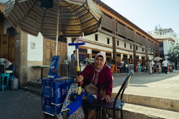 A woman wearing a colorful headscarf and a maroon sweater is seated next to a street vendor stand. She appears to be operating a manual machine, possibly an ice cream maker. There is a large striped umbrella providing shade. Behind her is a historic building with wooden doors and windows, creating a bustling outdoor market atmosphere. Several people are in the background, engaging in various activities.