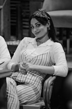 A person is seated on a chair, wearing a striped outfit with a matching headband. The individual has a pleasant expression, with hands clasped together. The setting appears to be indoors, possibly a casual or work environment.
