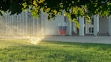 A sprinkler is watering the vibrant green grass under a canopy of trees, with sunlight glistening through the water droplets. In the background, a person rides a scooter near a building with numerous vertical columns, and a sculpture featuring elements of red, white, and blue stands prominently.